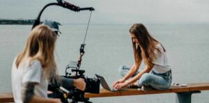 Εικόνα που δείχνει έναν καμεραμάν, ο οποίος κινηματογραφεί μια γυναίκα με ένα laptop την ώρα που εργάζεται, με φόντο τη θάλασσα. Η εικόνα αποτελεί στιγμιότυπο από τη δημιουργία βίντεο, το οποίο ανήκει στην κατηγορία: Social Media Video.