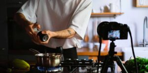 Εικόνα που δείχνει έναν μάγειρα καθώς μαγειρεύει και μια βιντεοκάμερα η οποία καταγράφει τη διαδικασία. Η εικόνα αποτελεί στιγμιότυπο από τη δημιουργία βίντεο, το οποίο ανήκει στην κατηγορία: Εκπαιδευτικά Βίντεο | Training video.